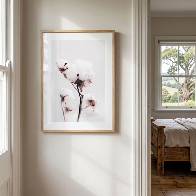 A vertical framed art print featuring white cotton bolls on a dark stem, displayed on a light beige wall in a bedroom setting.