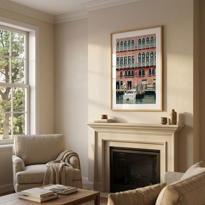 A framed art print depicting a canal-side building with arched windows, displayed on a beige wall above a fireplace in a styled living room with a chair, coffee table, and sofa.