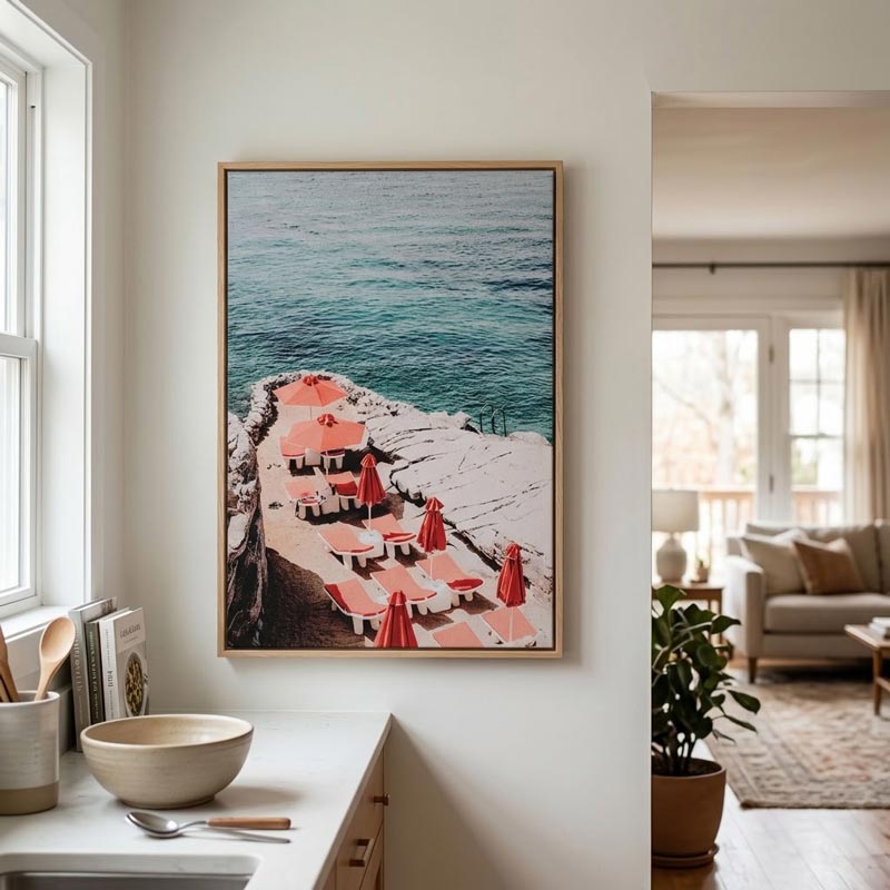A framed vertical art print depicting a coastal beach scene with turquoise water, white sand, and pink umbrellas, displayed on a white wall in a modern kitchen/living space.