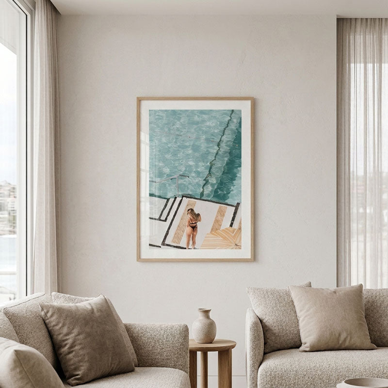 A framed art print featuring a minimalist illustration of a woman by a swimming pool, displayed on a light beige wall in a modern living room setting with two beige sofas and sheer curtains.