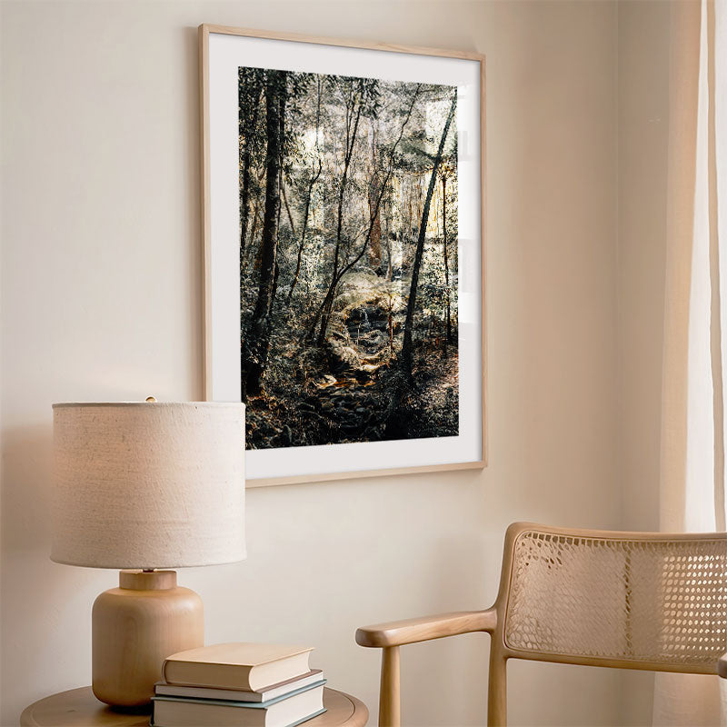 Framed forest print on a wall above a lamp and books with a chair to the side.