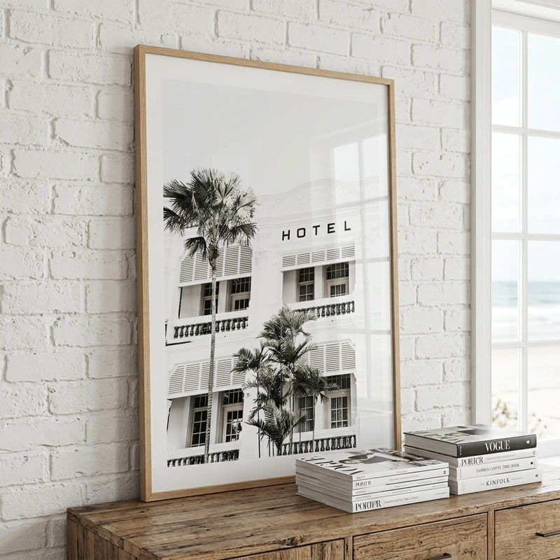 Framed black and white print of a hotel with palm trees on a wooden console table against a white brick wall.