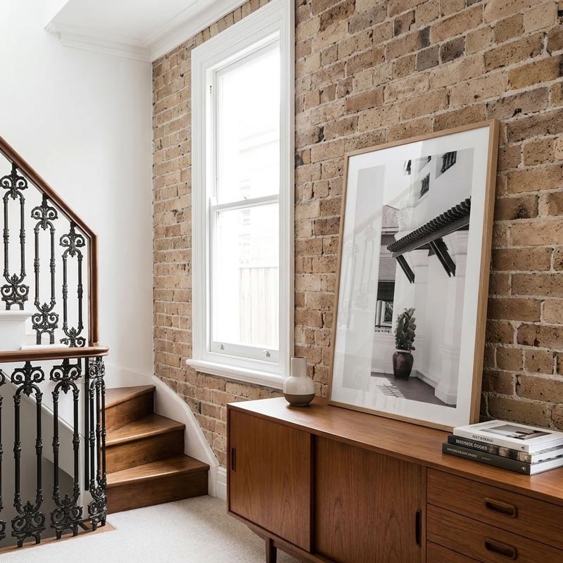 A large framed black and white architectural photograph leaning against a brick wall on a wooden credenza in a styled interior room.