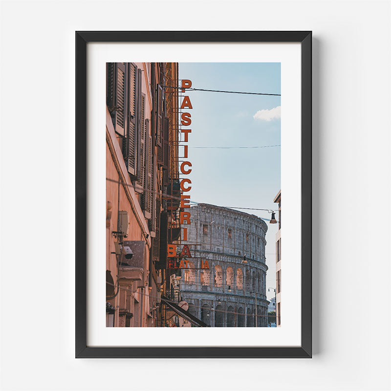 Framed wall art featuring a vertical photograph of the Colosseum in Rome with a tall red 'PASTICCERIA BIANCA' sign, shown in a black frame with a white mat against a light gray wall.