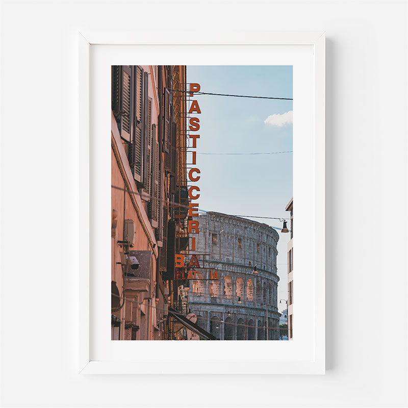 A framed art print featuring a photograph of the Colosseum in Rome with a vertical 'PASTICCERIA BIANCA' sign on a building, presented in a white frame against a light gray wall.