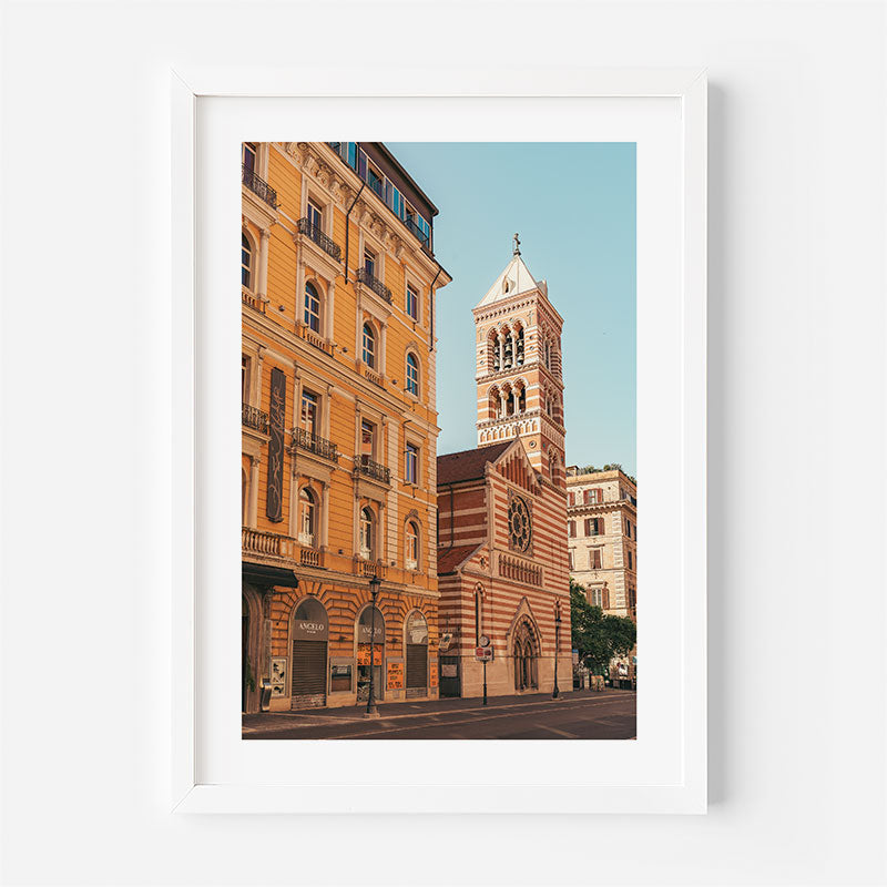 A framed art print featuring a scenic photograph of a European street with a church tower and warm-toned buildings, displayed on a clean white wall.