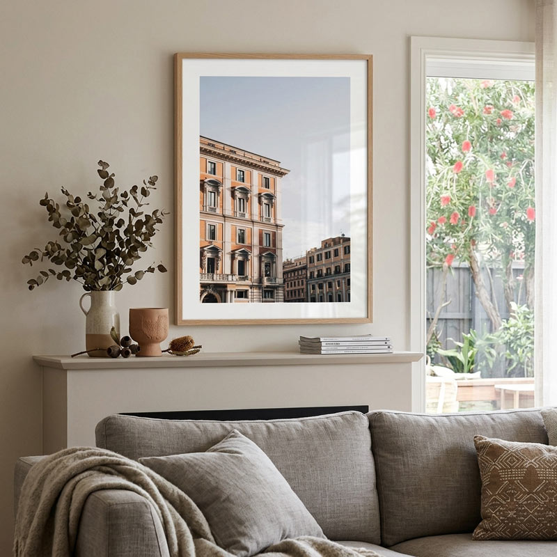 A framed art print depicting classic European buildings hangs on a beige wall above a gray sofa in a styled living room setting.
