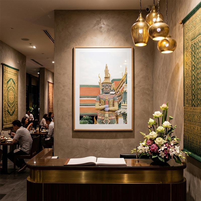 A framed art print depicting a Thai temple guard statue is displayed on a textured beige wall in a restaurant or cafe setting. The print is framed in a thin gold-toned frame and hangs above a dark wood reception desk with a gold trim, decorated with a white and pink flower arrangement.