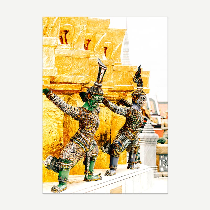 A vertical photographic print of two Thai garden statues in ornate green and gold armor, posed against a bright golden temple wall, shown centered on a light gray background.