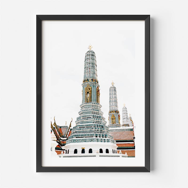 A framed art print depicting the ornate spires of Wat Arun in Bangkok, featuring a white and gold temple structure against a pale sky, presented in a black frame with a white mat.