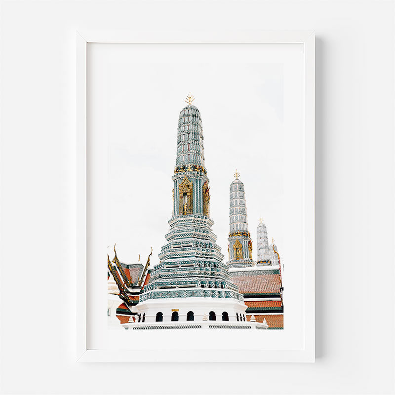 A framed art print depicting the ornate, tiered spires of Wat Arun in Bangkok, featuring white, gold, and blue-gray tones, presented in a clean white frame against a white wall.
