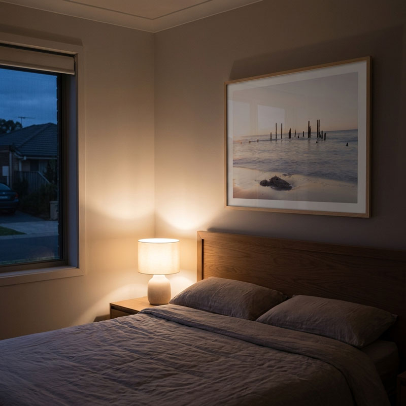 A styled bedroom scene with a neatly made bed, bedside lamp, window view at dusk, and a framed beach photograph on the wall.