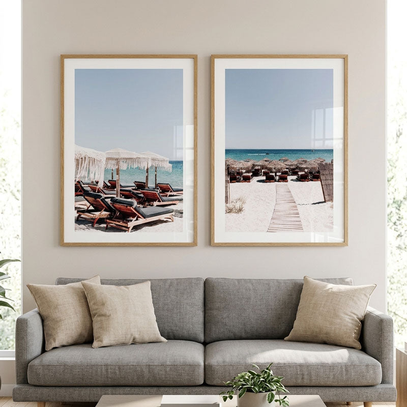 Two vertical framed beach prints hanging side by side on a light beige wall above a gray sofa with beige pillows in a bright living room.