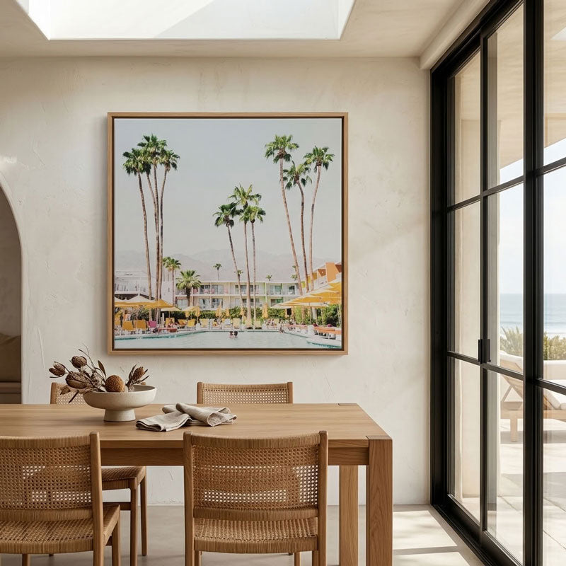 A framed art print depicting a sunny poolside resort scene with tall palm trees is displayed on a beige wall above a wooden dining table with woven chairs in a modern interior.