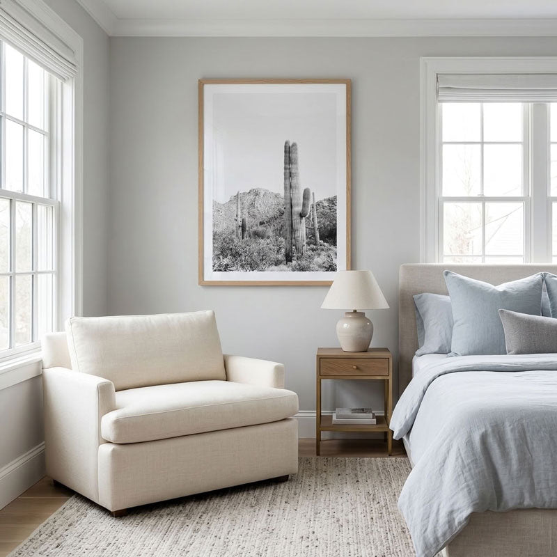 A framed black and white desert cactus art print hangs on a light gray wall in a styled bedroom, above a cream armchair and next to a bed with light blue bedding.