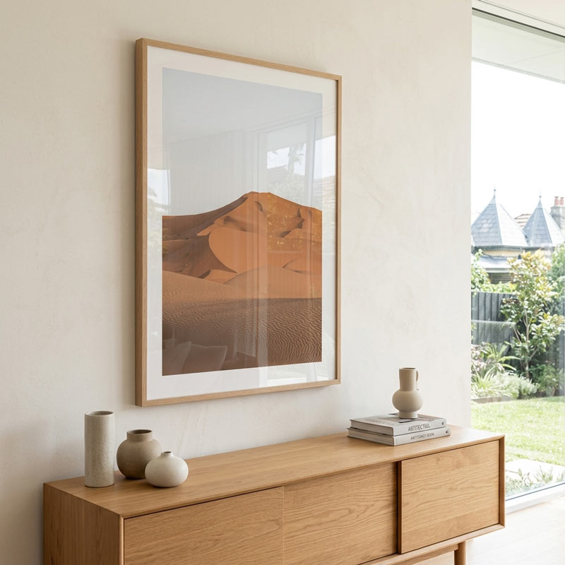 A large framed art print depicting desert sand dunes in warm brown and orange tones, styled on a cream wall above a light wood console table with neutral ceramic vases.