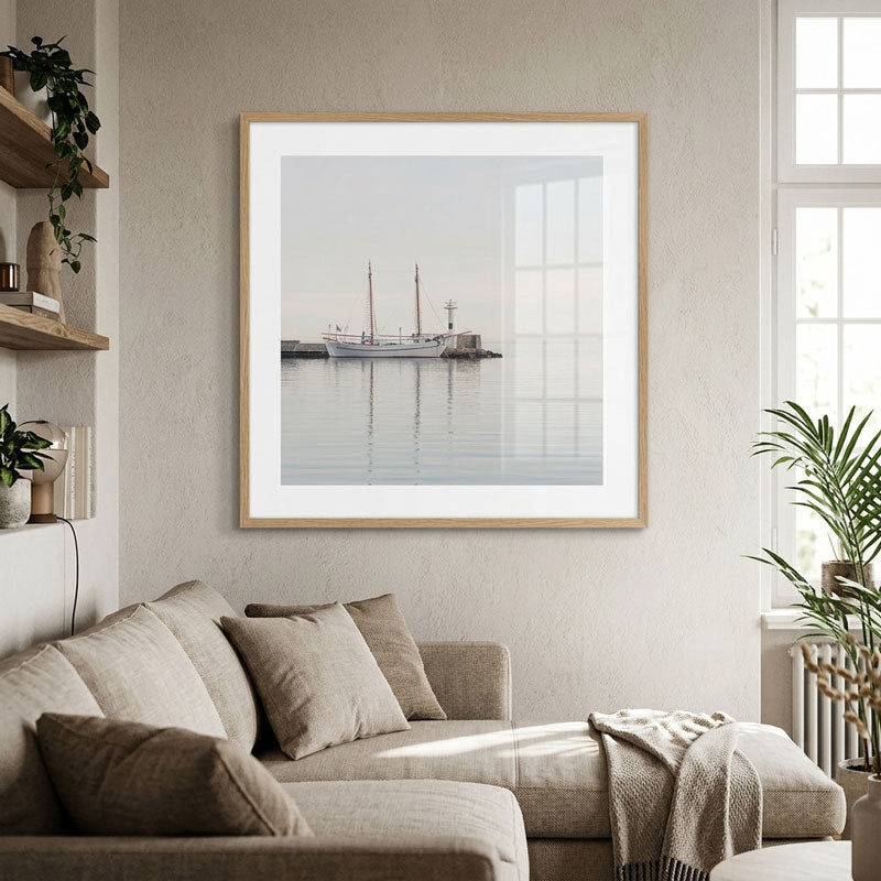 A framed art print depicting a calm harbor scene with a sailboat and piers, styled on a beige wall above a modern beige sofa in a bright, contemporary living room.