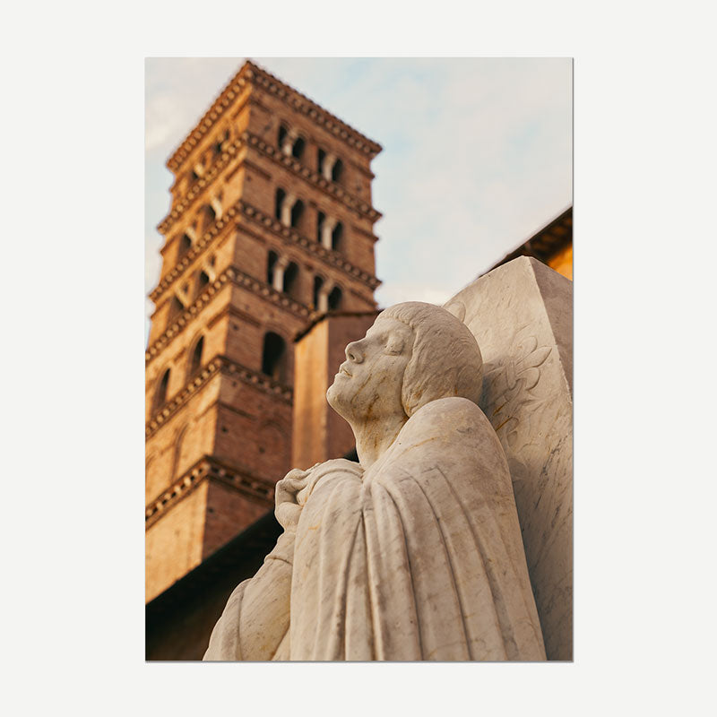 A vertical photographic print or canvas wall art featuring a close-up of a stone angel statue in the foreground with a warm-toned brick bell tower and pale sky in the background.