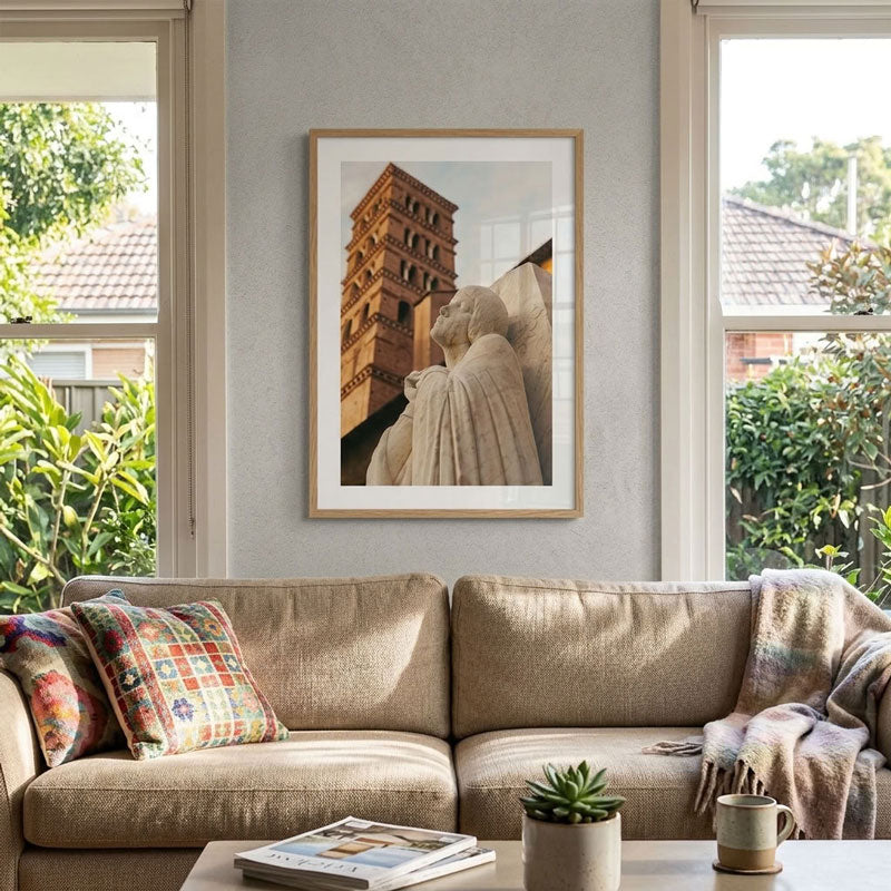 A framed art print depicting a stone statue and a brick tower, displayed on a light gray wall above a beige fabric sofa in a styled living room.