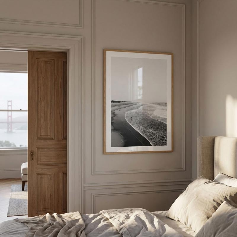 A framed black and white landscape photograph of a beach shoreline, styled in a light wood frame and hung on a beige paneled wall above a bed in a modern bedroom.