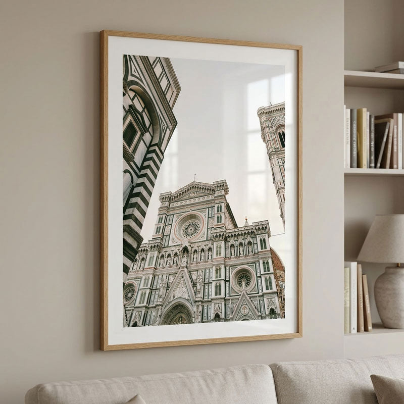 A framed art print of the Florence Cathedral (Santa Maria del Fiore) hanging on a beige wall in a living room setting, next to a bookshelf with books and a lamp.
