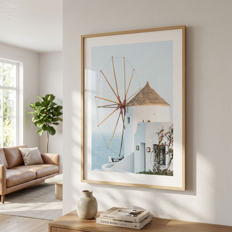 A framed art print depicting a white building with a windmill and a thatched roof against a light blue sky, hanging on a white wall in a modern, sunlit living room.