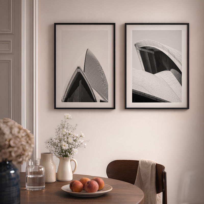 Two framed black and white artworks on a wall above a dining table with fruit and flowers.