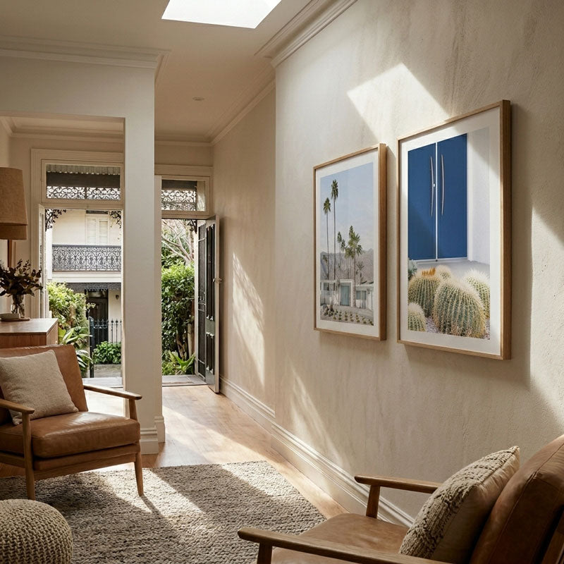 Two framed art prints hang on a beige wall in a sunlit living room. The left print shows palm trees and a house, and the right print shows cacti with a blue cabinet.