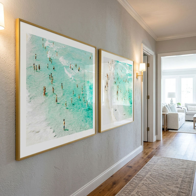 Two large framed art prints depicting turquoise ocean waves with small figures, hung on a light gray wall in a modern hallway with wood floors.