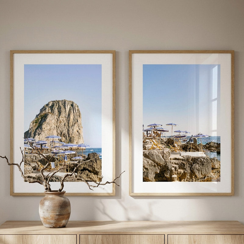 Two framed beachscape prints on a wall with a vase and branches in the foreground.