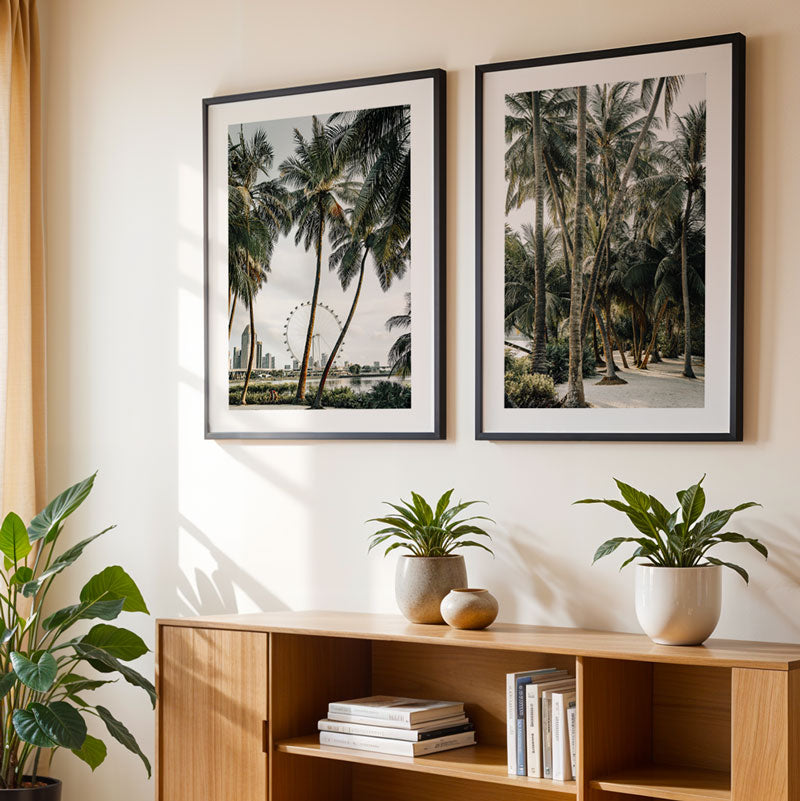 Two framed palm tree prints on a wall above a wooden shelf with books and plants.