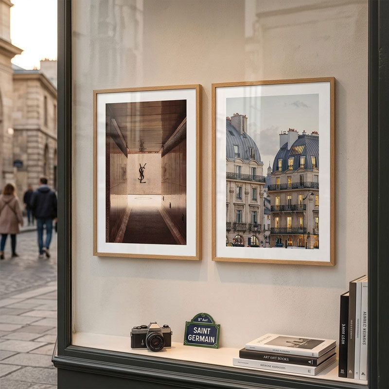 Two framed art prints displayed side-by-side on a beige wall inside a window or storefront. The left print is a sepia-toned photograph of a person in a tunnel, and the right print shows a Parisian street scene with classic buildings.