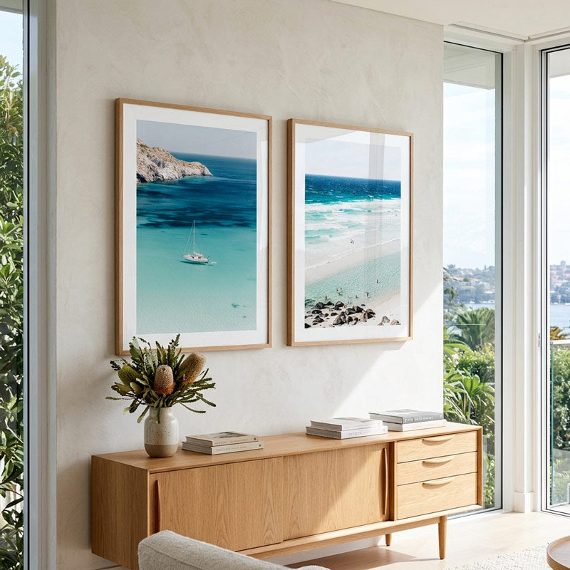 Two framed beach and sea art prints hang on a light beige wall above a wooden sideboard in a bright, modern room. The prints show turquoise ocean waters, rocky coastline, and a sandy beach.