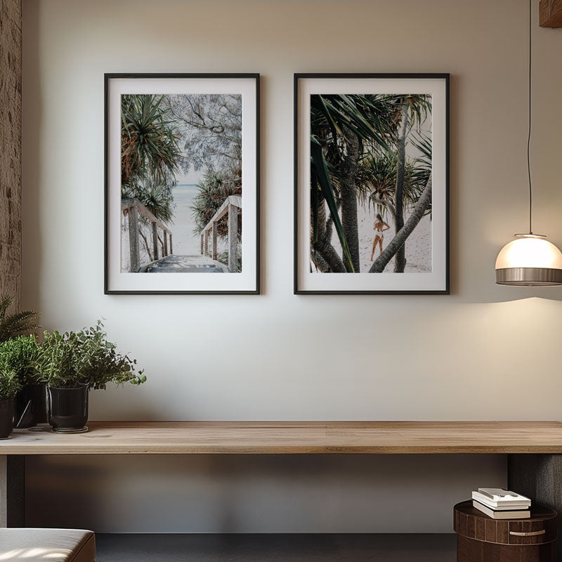 Two framed artworks on a wall above a wooden bench with a lamp and plants.