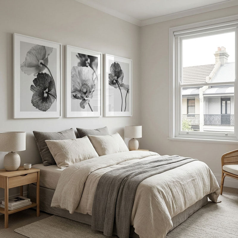 Neatly made bed with neutral bedding in a bedroom with floral wall art and a window.