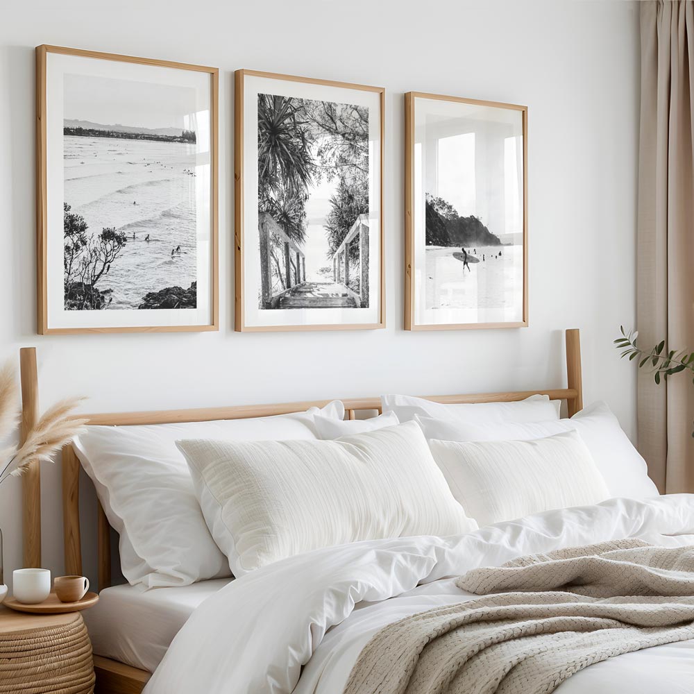 A set of three black and white framed photographs displayed above a bed, depicting surfers and coastal scenes.