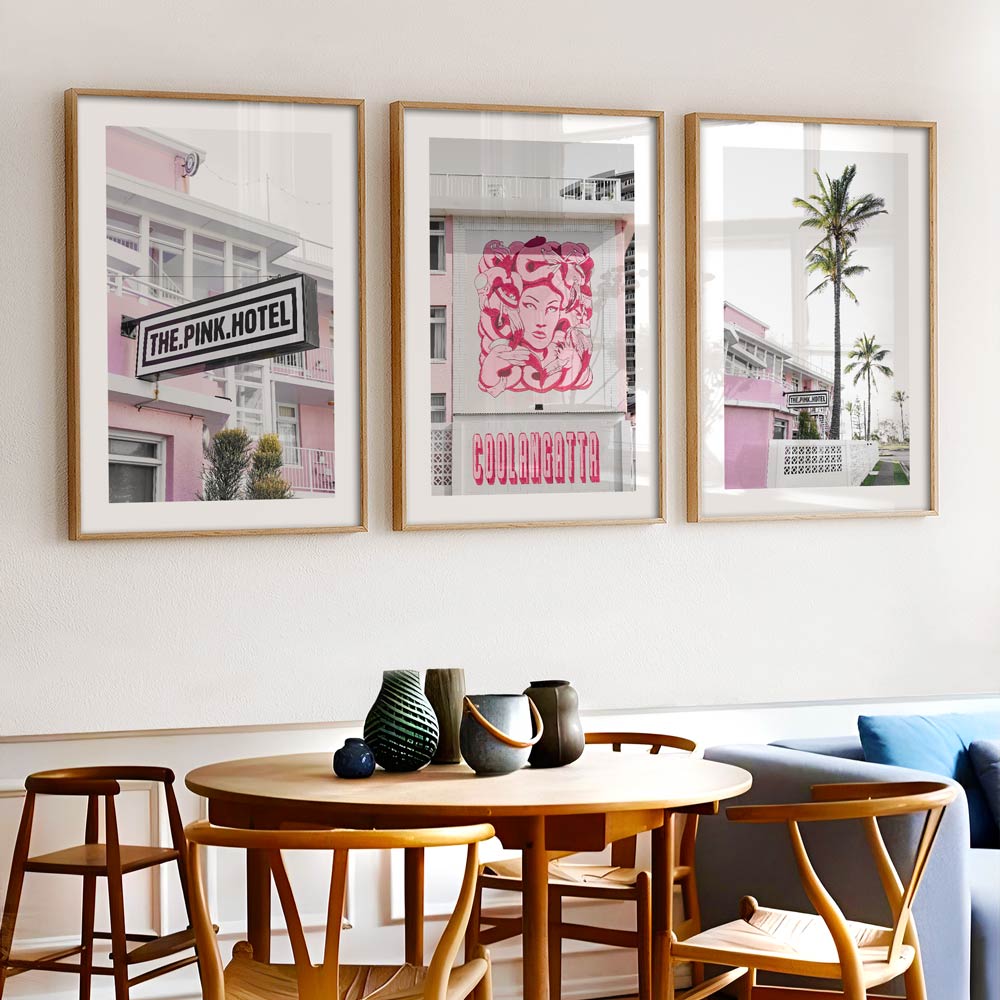 A set of three framed photographs featuring 'The Pink Hotel' displayed on a wall above a dining area with a round wooden table and modern chairs.