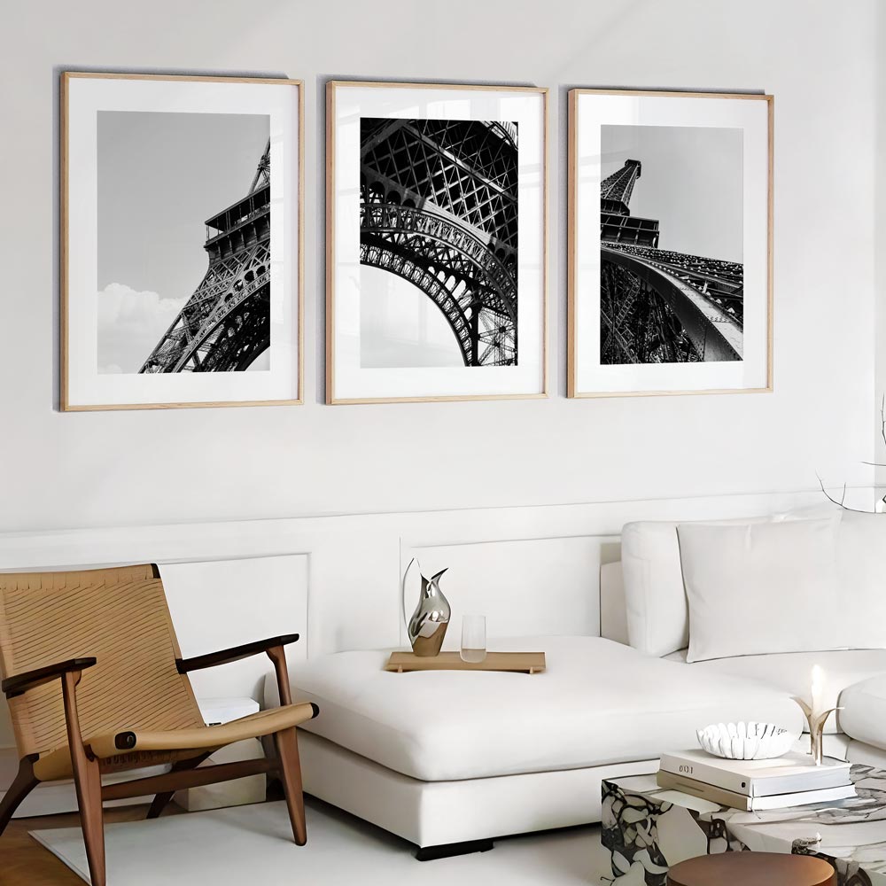A set of three black and white photographs in frames, depicting the Eiffel Tower, placed above a white sofa in a room with a minimalist decor.