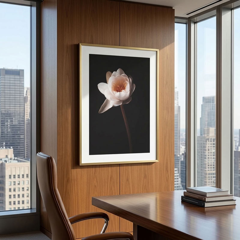 A framed wall art print featuring a pale pink flower on a dark background, displayed in a modern office setting with wood paneling, a desk, and city windows.