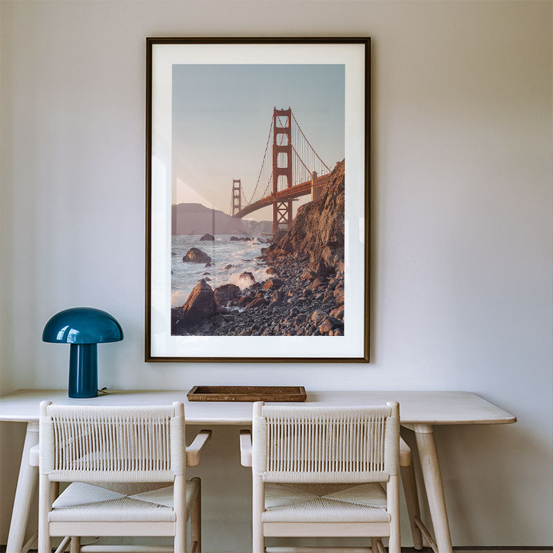 Framed photograph of the Golden Gate Bridge above water on a wall above a dining table with chairs.