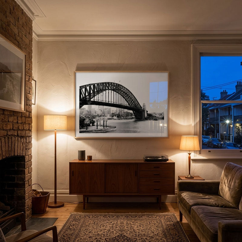 A large framed black and white art print of the Sydney Harbour Bridge hangs on a beige wall in a beautifully styled living room with a wooden sideboard, lamps, a brick fireplace, and a sofa.
