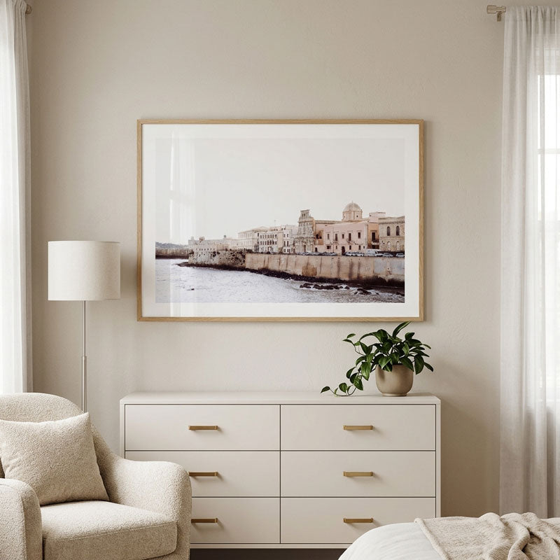 A framed art print depicting a coastal cityscape with historic buildings and water, displayed on a beige wall above a white dresser in a styled bedroom setting.