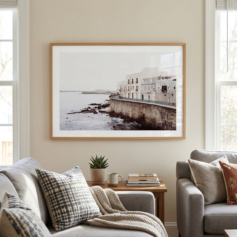A framed coastal art print hangs on a beige wall in a modern living room setting, showing a stone seaside building and rocky shoreline. The room includes gray upholstered chairs, patterned pillows, a small wooden side table with books and a plant, and bright windows on both sides.