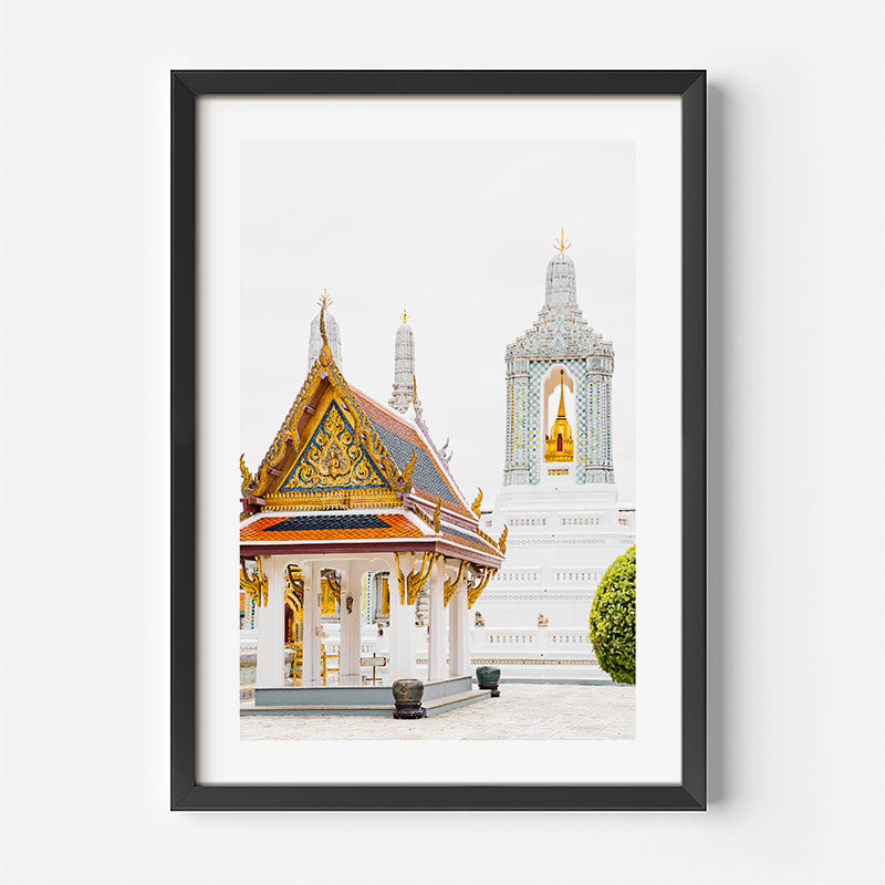 A framed art print of a Thai temple courtyard, featuring a golden-roofed pavilion and a white temple tower with a golden Buddha statue, presented in a black frame with a white mat against a light gray wall.