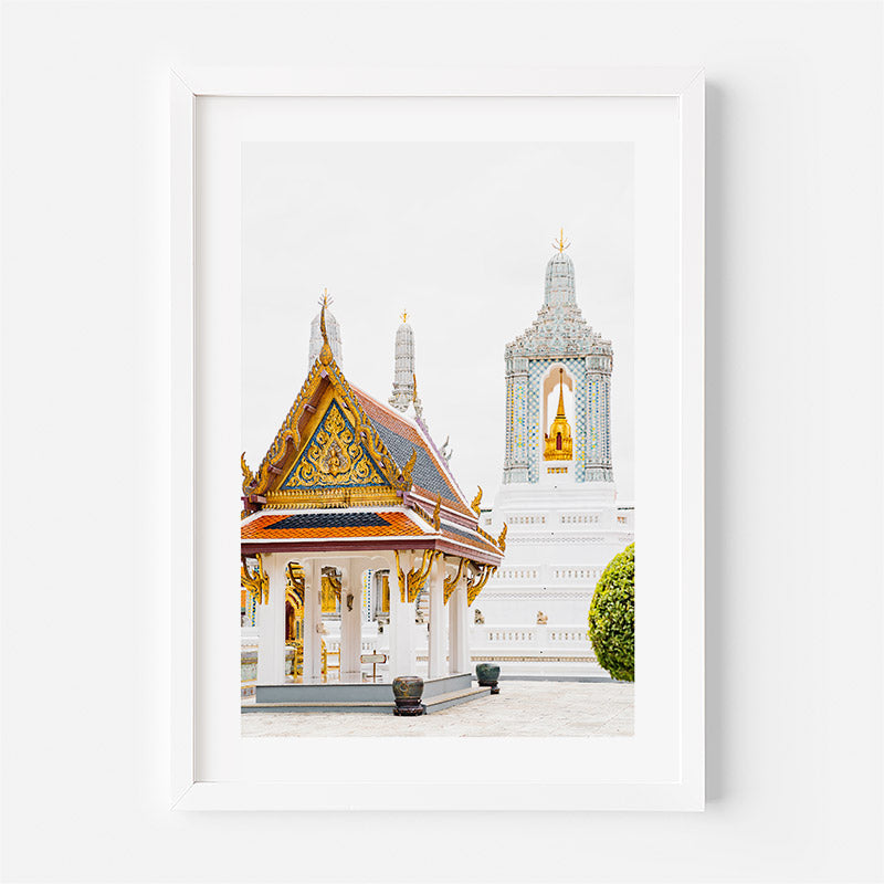 A framed art print depicting a Thai temple pavilion with a golden roof and a white temple tower in the background, presented in a white frame against a light gray wall.