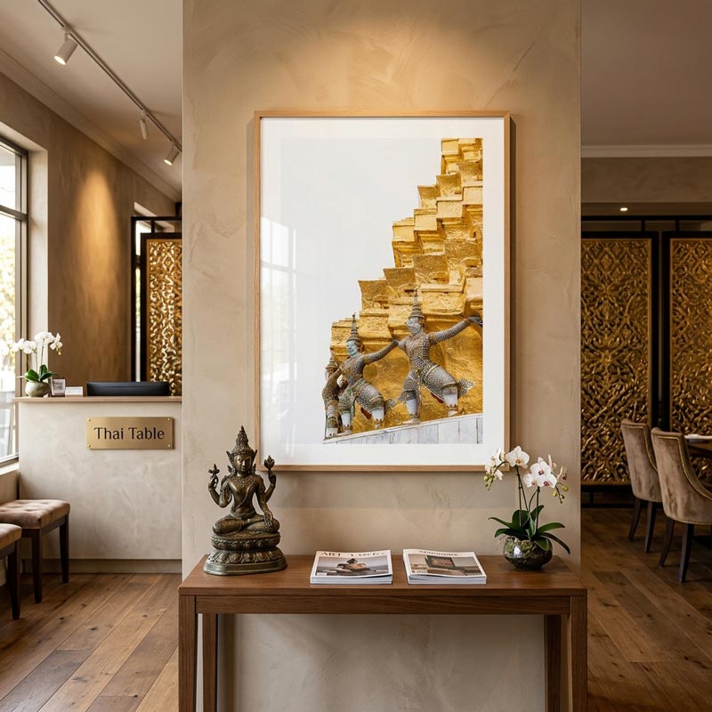 A framed art print depicting Thai temple dancers against a golden stepped background, displayed on a beige wall in a modern, stylishly decorated interior with a wooden console table and decorative statues.