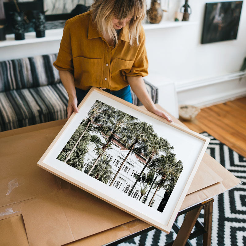 Woman holding a framed photo of trees and buildings in a living room.