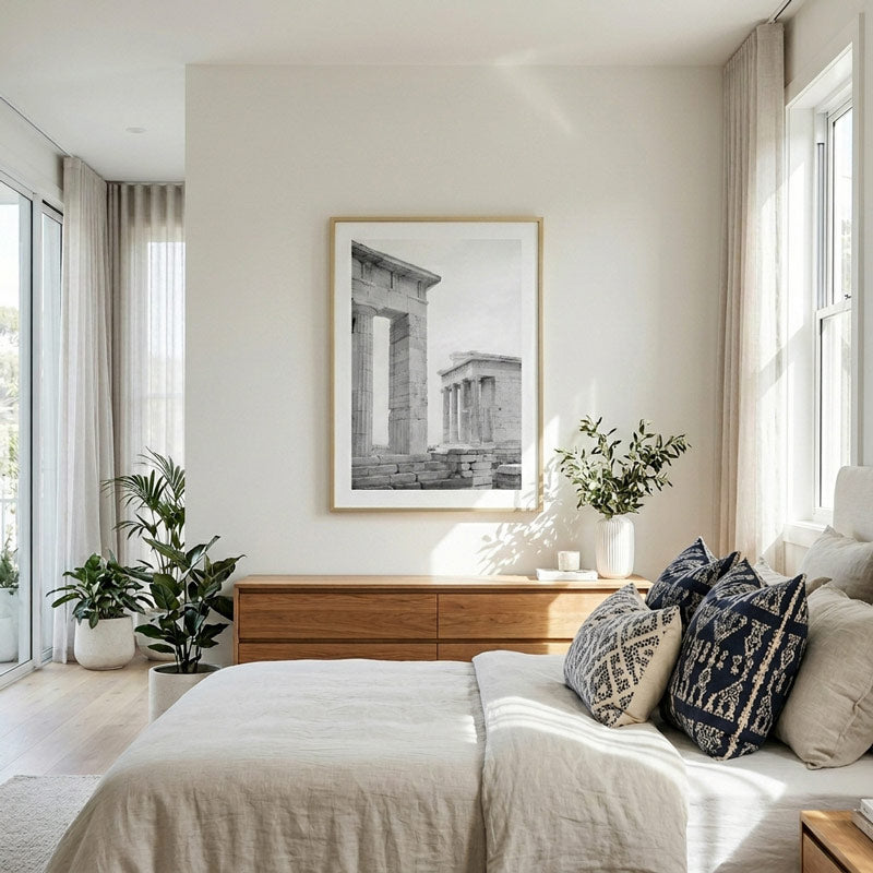 A framed black and white architectural art print of ancient ruins hangs on a warm off-white bedroom wall above a wooden dresser, flanked by plants and a neatly made bed with neutral-toned linens and patterned pillows.