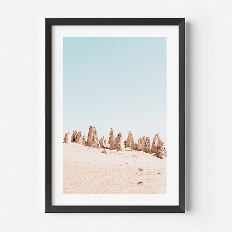 Framed artwork of desert landscape with rock formations against a light blue sky.