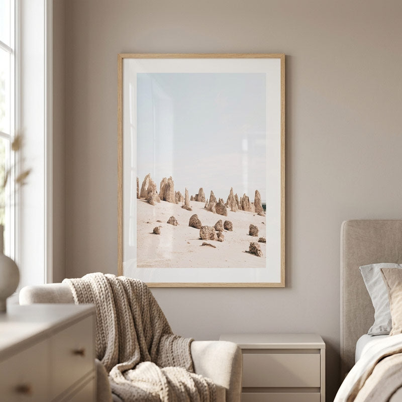 A framed art print depicting a desert landscape with rock spires under a pale sky, styled on a beige wall above a bed and next to a window in a bedroom setting.
